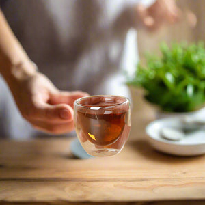 Hand holding a small glass cup of tea on a wooden table with two more cups in the background.